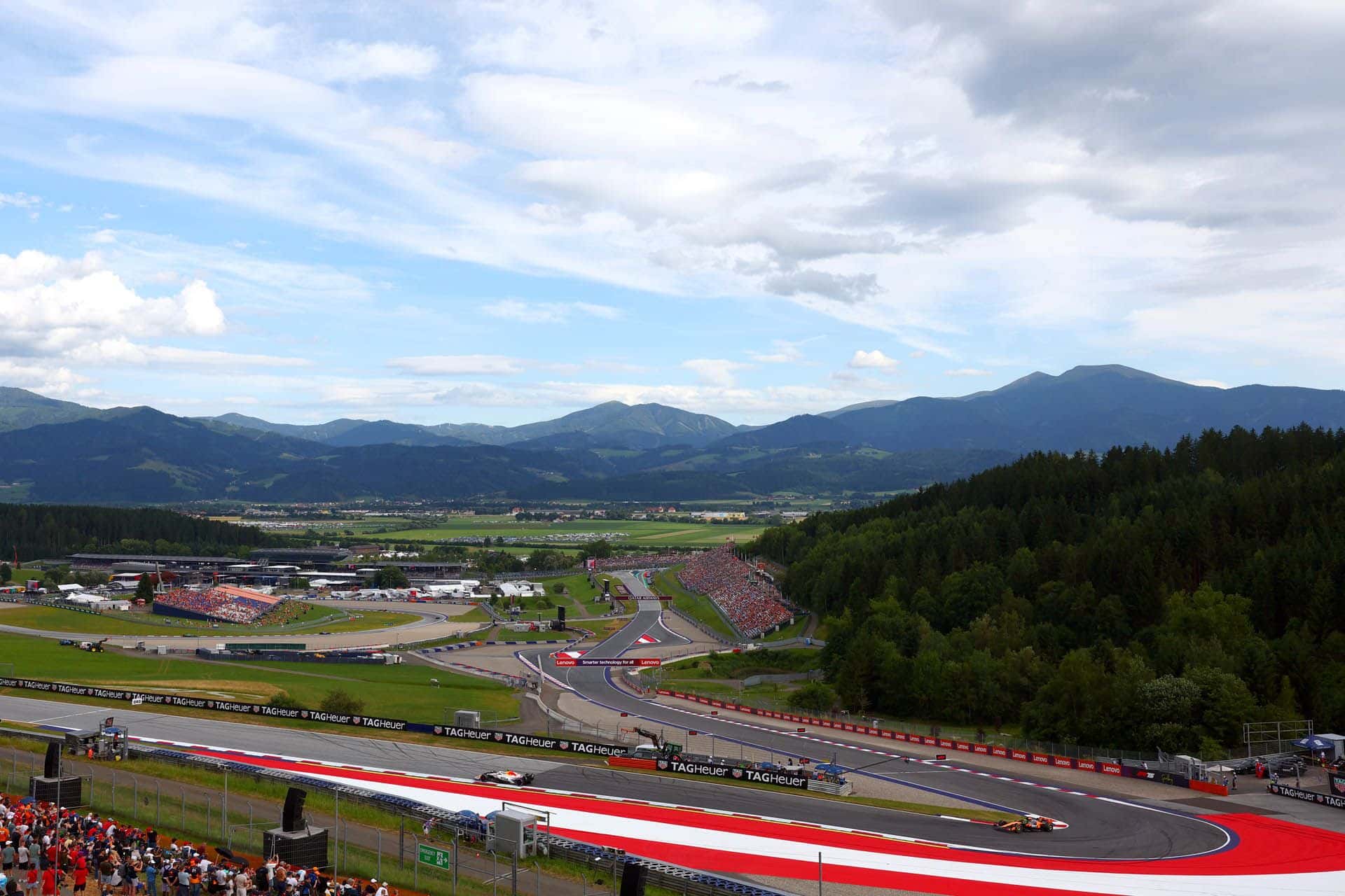F1 cars sweeping through the final corners at the Red Bull Ring, with packed grandstands and the Styrian mountains beyond