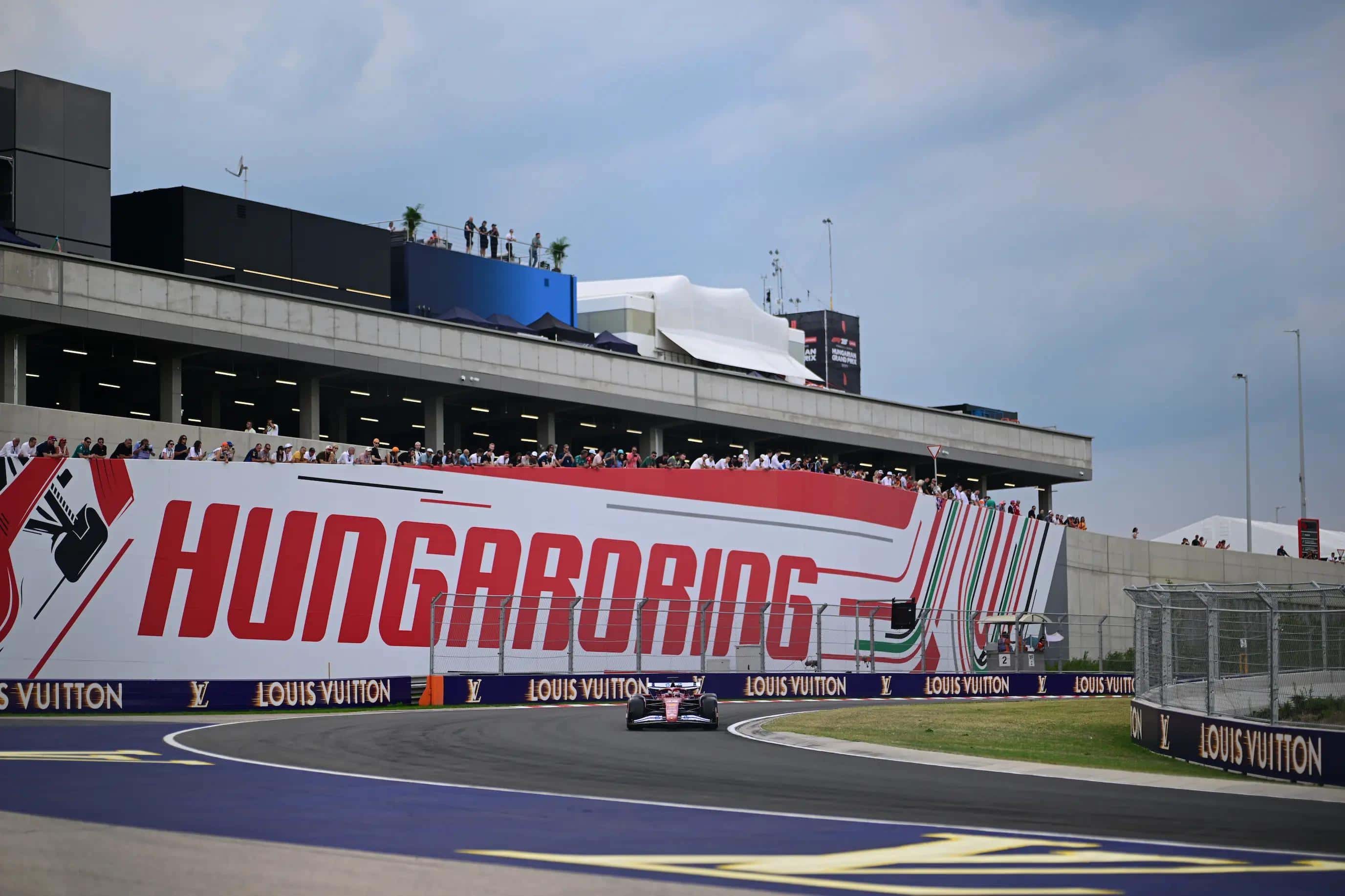 An F1 car passes the giant Hungaroring branding in front of the main grandstand complex