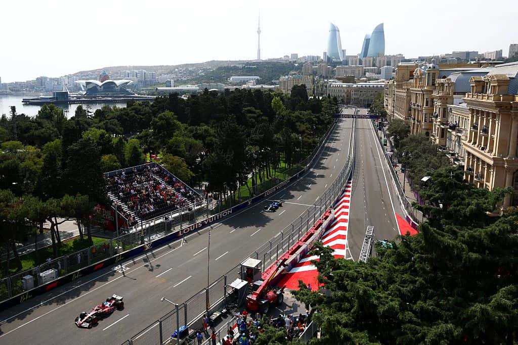 F1 cars on the tree-lined main straight of the Baku City Circuit, with the Flame Towers and Caspian shoreline in the distance