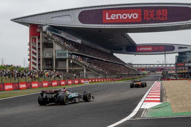F1 cars exiting the final corner at Shanghai International Circuit beneath the sweeping main grandstand roof